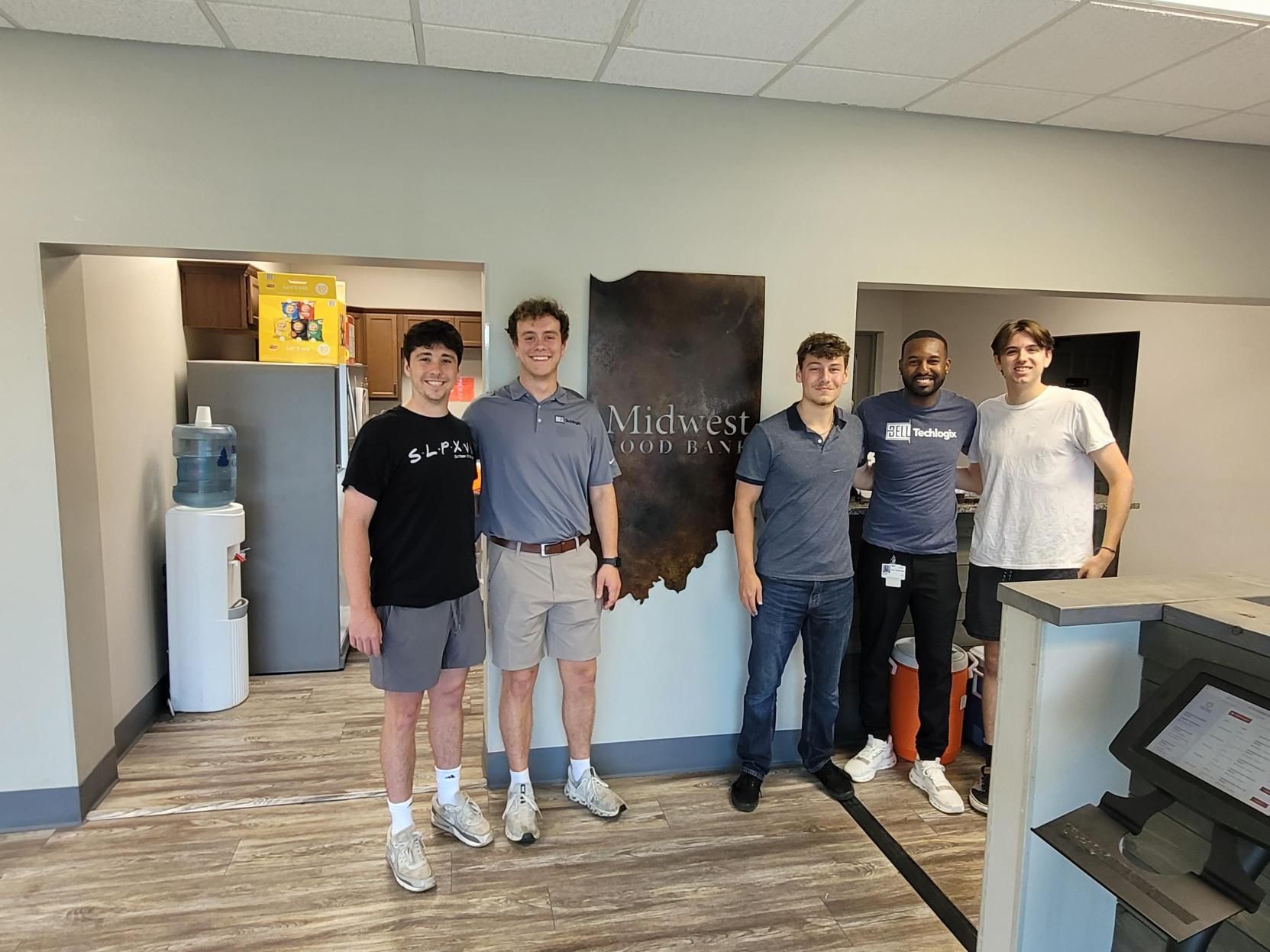 Seven Bell Techlogix summer interns and staff posing together in front of the Bell Techlogix wall sign in the office lobby