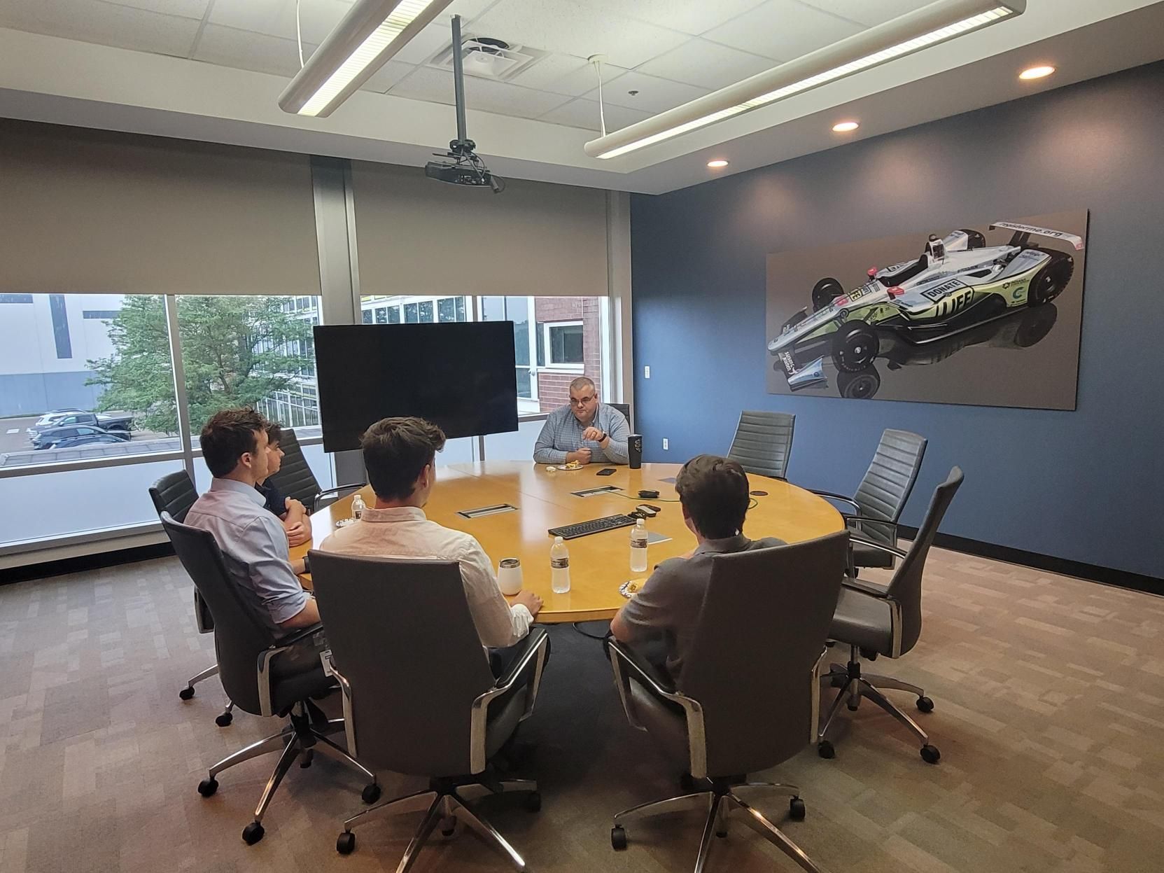 Bell Techlogix staff gathered around a conference table for a team meeting, with an IndyCar painting on the wall behind them