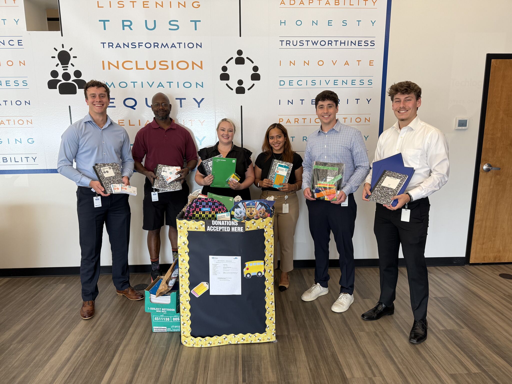Six Bell Techlogix team members holding donated school supplies — notebooks, folders, pencils — next to a decorated donation box, standing in front of a company values wall