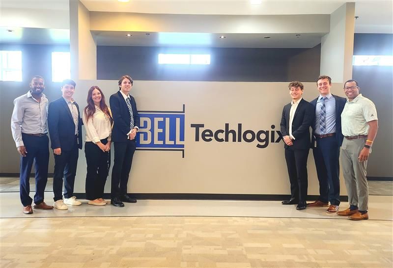 Seven Bell Techlogix summer interns and staff standing together in front of the Bell Techlogix wall sign in the office lobby