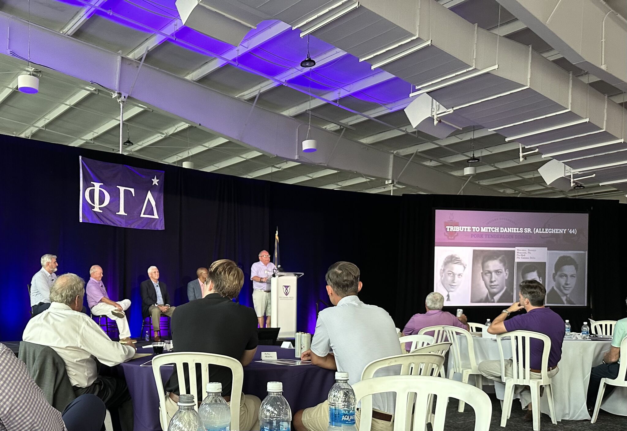 A panel discussion on stage, with a tribute slide honoring Mitch Daniels Sr. (Allegheny '44) displayed on the screen behind the panelists