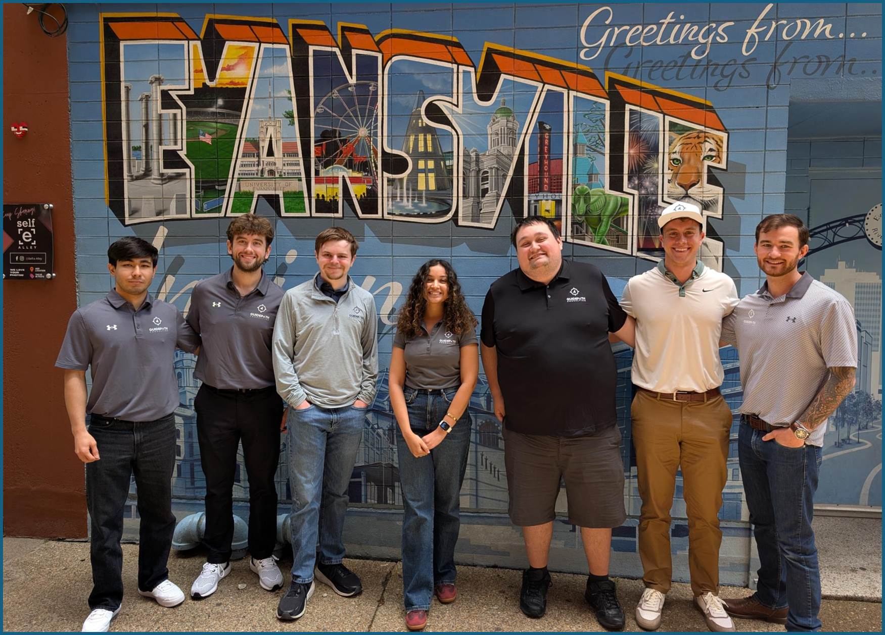 The Guidepath Consulting team standing in a row in front of the "Greetings from Evansville" mural in downtown Evansville, most wearing Guidepath polo shirts