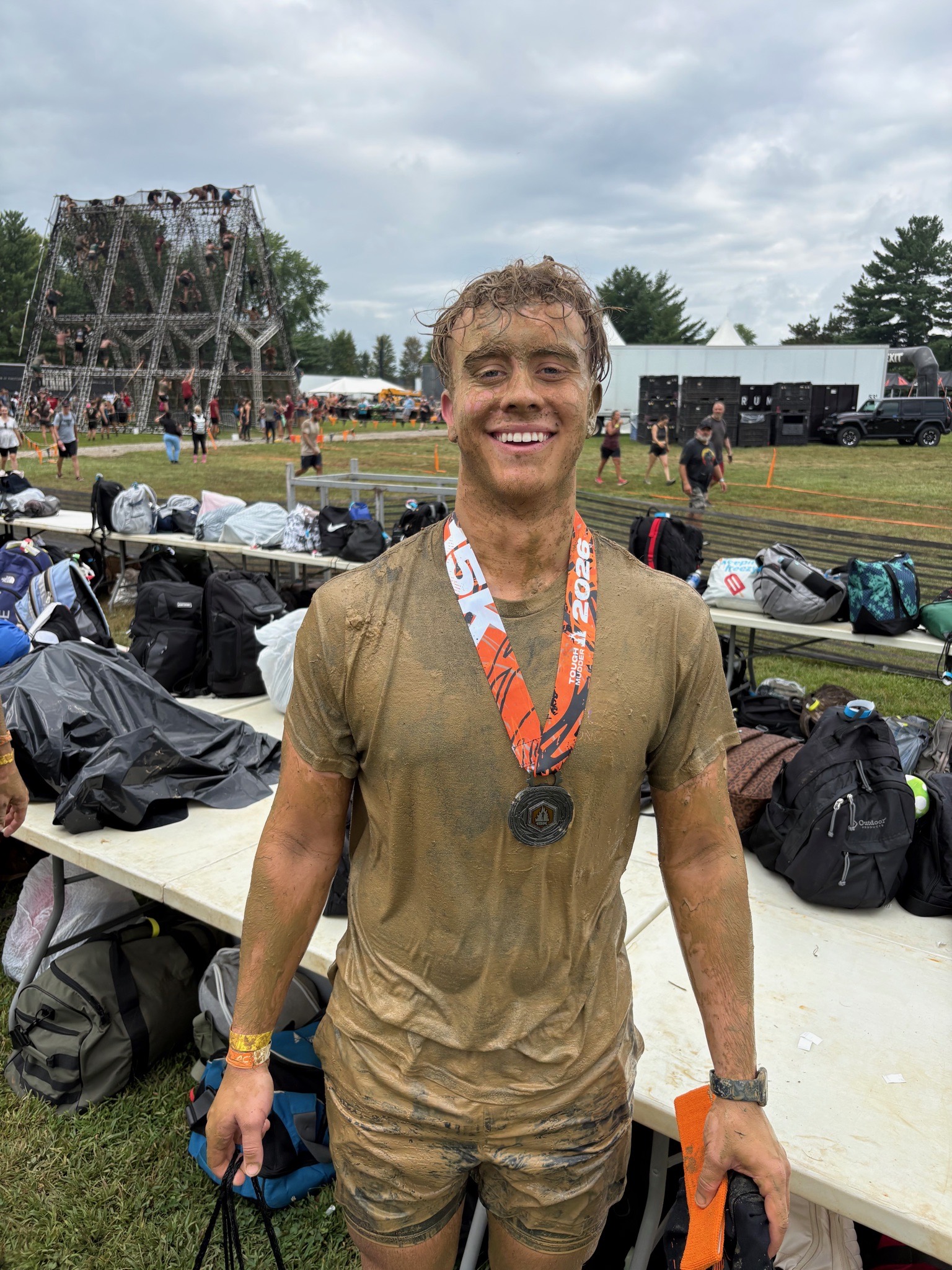 Covered in dried mud at the bag drop after finishing, wearing the 2026 Tough Mudder finisher medal
