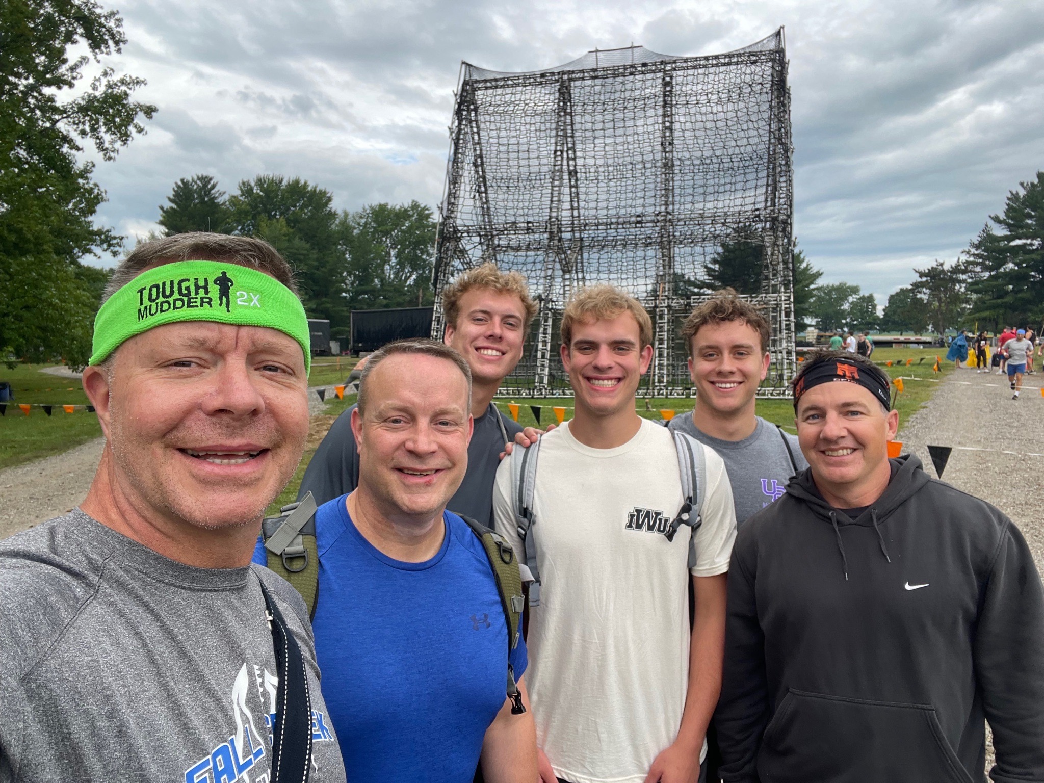 Our team posing together on the gravel path before the race, clean and dry, in front of the tall netted climbing obstacle