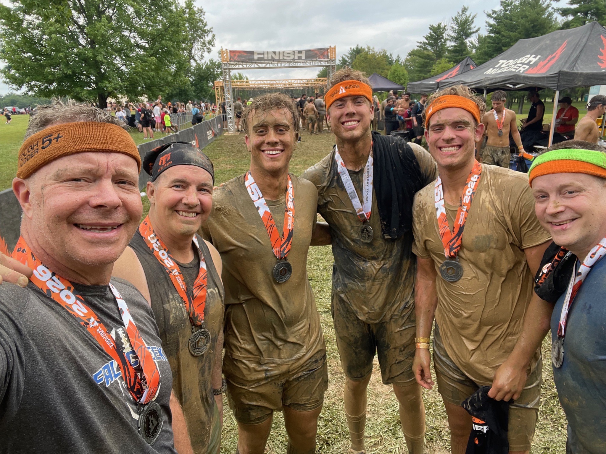 Six mud-covered Tough Mudder finishers, including Andrew S. Thompson, posing arm in arm with their medals in front of the finish line arch